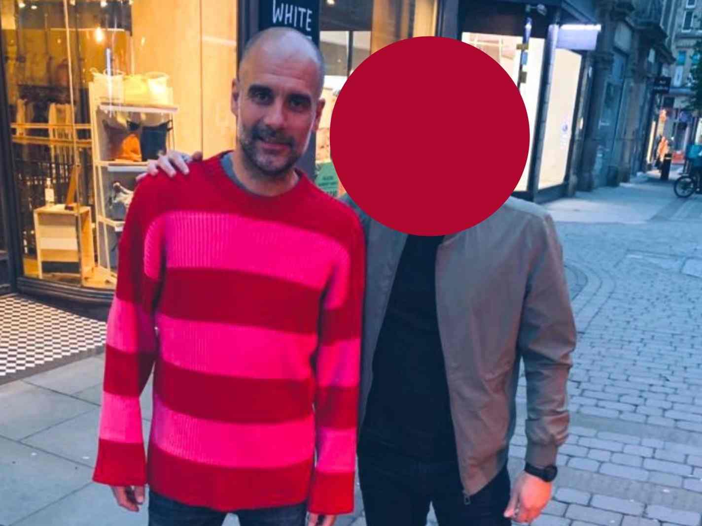 The image shows Pep Guardiola wearing a bright pinkred Balenciaga sweater and getting a picture take with a fan, whose face is blurred.